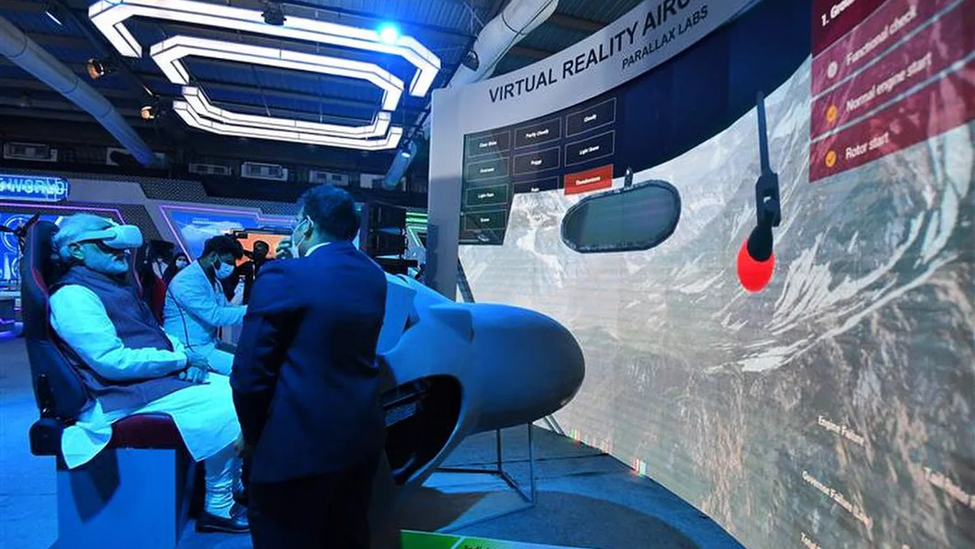 The Prime Minister of India in the simulator seat at Defense Expo 2022, headset on, an operator beside him and the terrain running on the curved projection wall behind